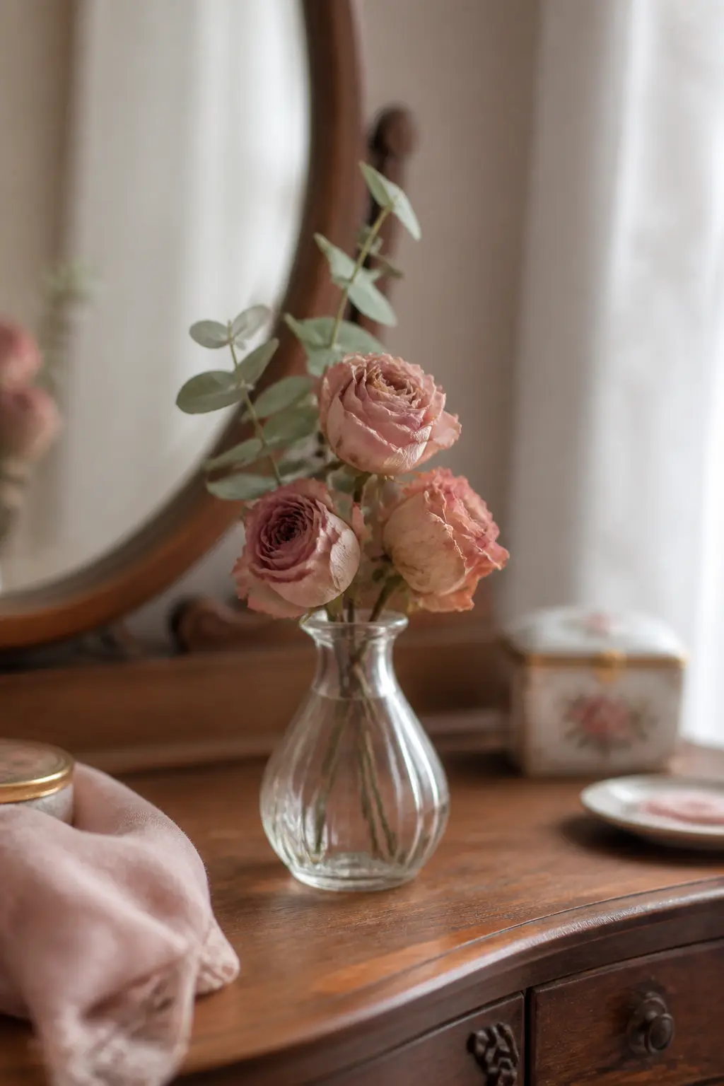 A clear glass bud vase of soft dusty-rose dried roses and eucalyptus on a wooden vanity in soft daylight.